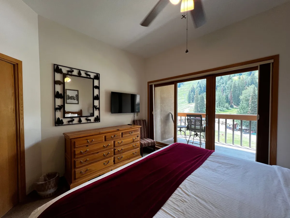 This image features a queen-sized bed adorned with a burgundy blanket, a wooden dresser for ample storage, a large window that frames beautiful natural views, and a TV mounted on the wall. The scene exudes tranquility and comfort, making it an ideal representation of a restful bedroom retreat.