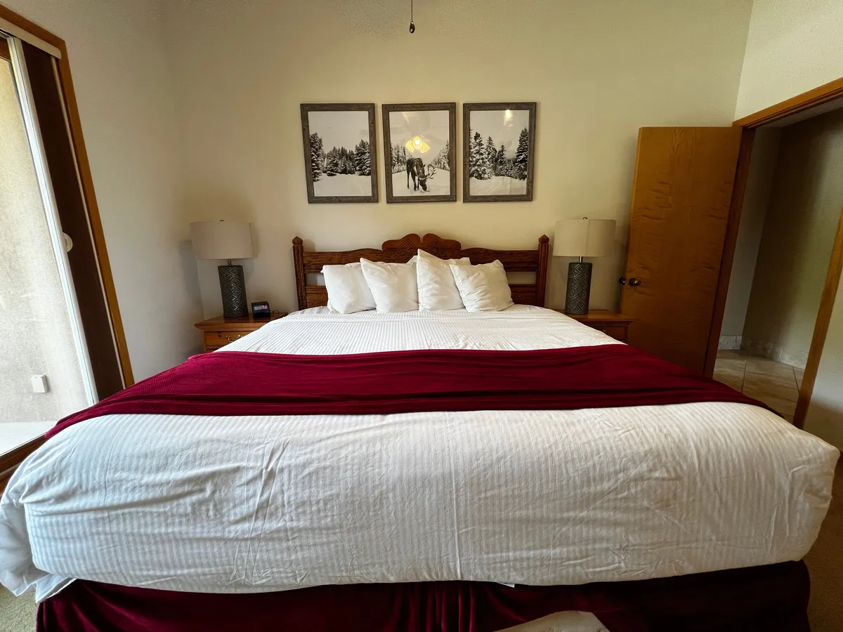 A spacious bedroom highlighted by a large bed with crisp white bedding and a warm red throw, complemented by three framed pictures on the wall and a cozy nightstand with a lamp. The lighting is soft and inviting, creating a peaceful ambiance ideal for relaxation.