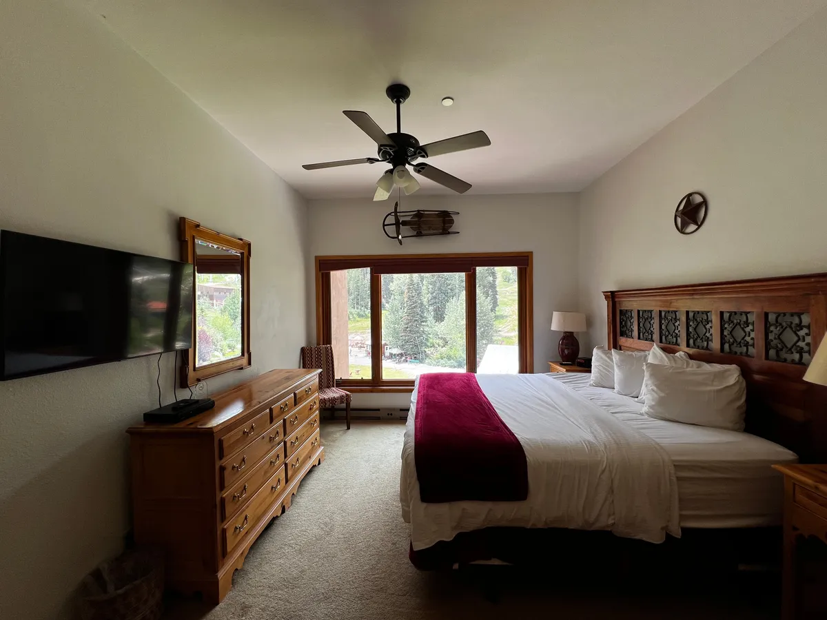 This serene bedroom showcases a large window with a forest view, a sturdy wooden bed frame, a dresser with a mirror, and a ceiling fan. The scene encapsulates the feeling of waking up surrounded by nature and enjoying calm, restorative moments indoors.
