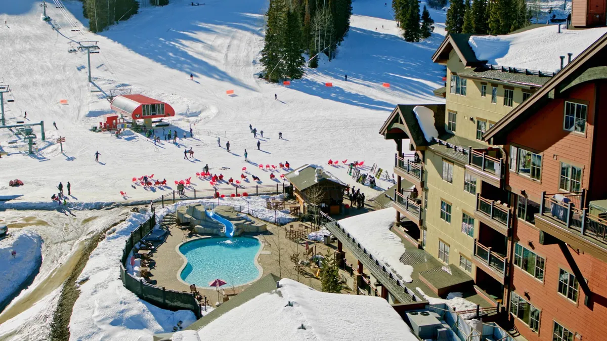 An outdoor pool area adjacent to a ski lodge, with snow-covered slopes, a ski lift, and guests enjoying both swimming and winter sports. The lodge building appears welcoming, and the juxtaposition of the pool with ski activities emphasizes year-round amenities.