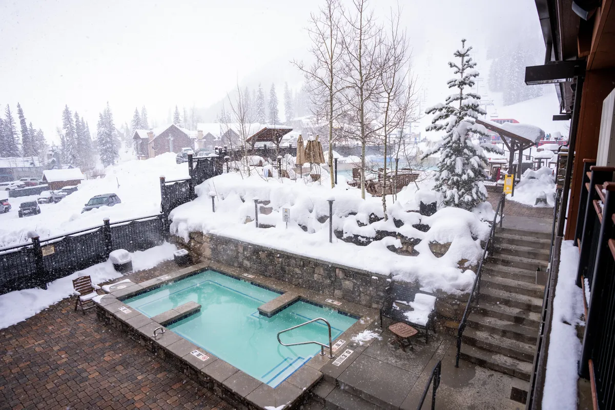 A rooftop hot tub nestled amidst snow-covered trees with a breathtaking view of the surrounding mountains and a snowy landscape. People are relaxing in the warm waters while the winter scenery unfolds around them, suggesting an inviting spot for stargazing or socializing after a day on the slopes.
