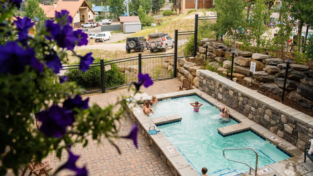 An inviting outdoor hot tub and swimming pool framed by flowers and mountain scenery, with residential buildings in the background. The scene radiates relaxation and social enjoyment after a day on the slopes.