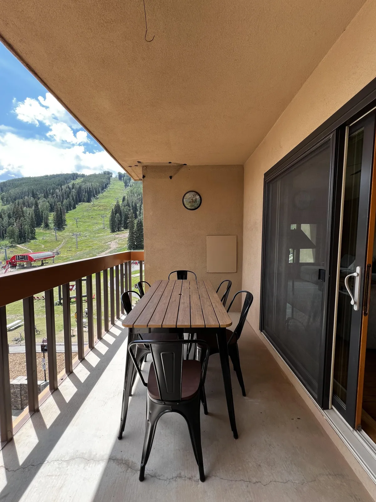 An exterior patio with a wooden table and metal chairs set against a dramatic mountain view. The clock on the wall suggests relaxed moments spent outdoors, while the setting is ideal for socializing, dining, or enjoying a morning coffee with an alpine backdrop.