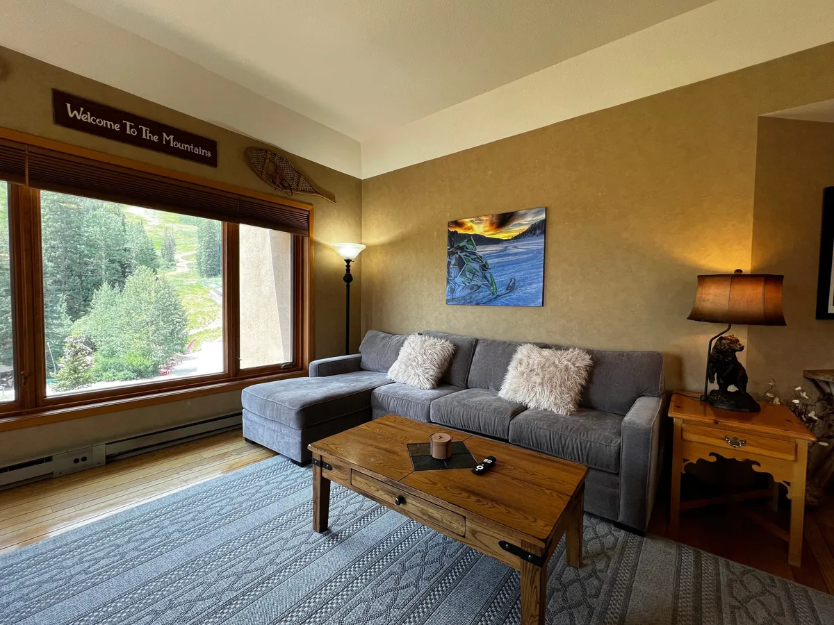 A bright, inviting living space with a cushioned sofa, wooden coffee table, and a wide window looking out onto the majestic mountains. The room glows with the golden light of sunset, enhancing the sense of warmth and comfort ideal for relaxing after a day on the slopes.