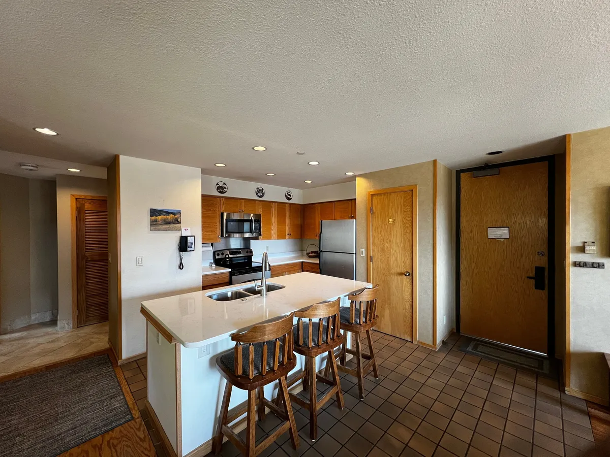 This interior shot centers on a kitchen island with bar stools, modern appliances, and a glimpse of the entrance door. The arrangement encourages interaction, with space for guests to gather around the island for casual meals, coffee, or conversation. The overall mood is inviting and conducive to both quick breakfasts and leisurely gatherings.
