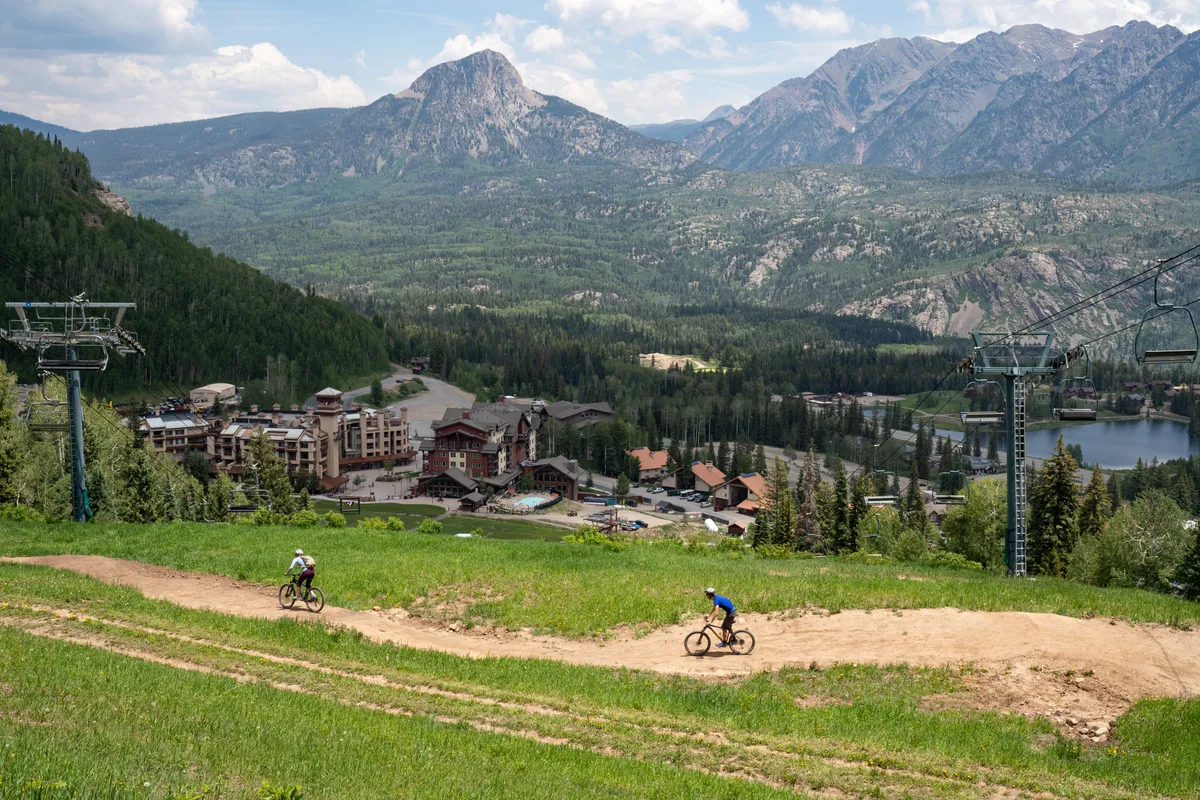 A panoramic exterior view showing Purgatory Resort’s dramatic mountain landscape. Mountain bikers and hikers are visible, with resort buildings and a ski lift in the background, and a sparkling lake nearby. The scene is lively and full of outdoor adventure possibilities.