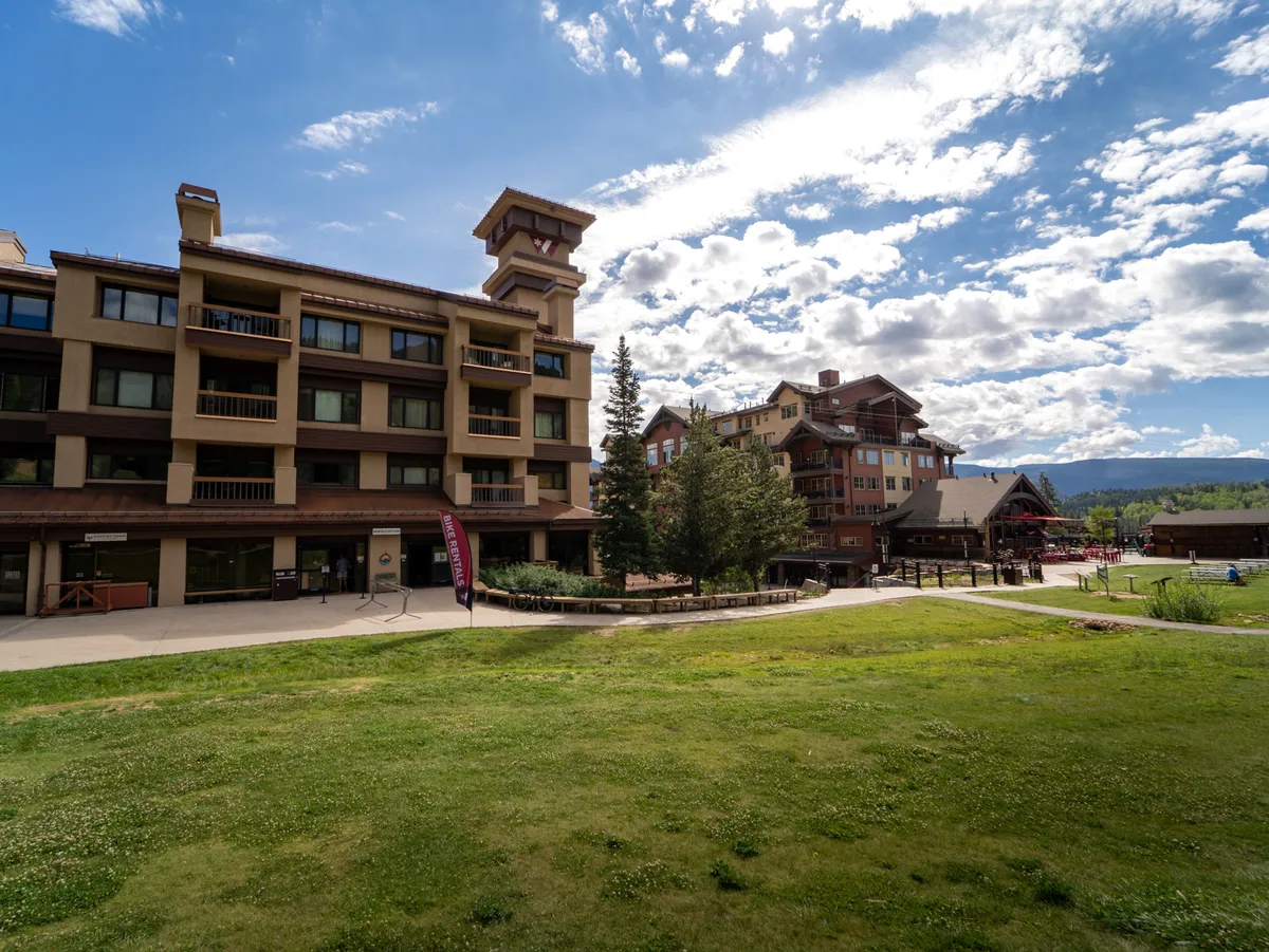 This image shows the main building of Village Center nestled amidst a landscaped garden, with the majestic San Juan Mountains rising in the background. The exterior is welcoming, and the setting highlights the property's close connection to the alpine environment. Sunny weather and lush greenery suggest a vibrant, inviting resort atmosphere.