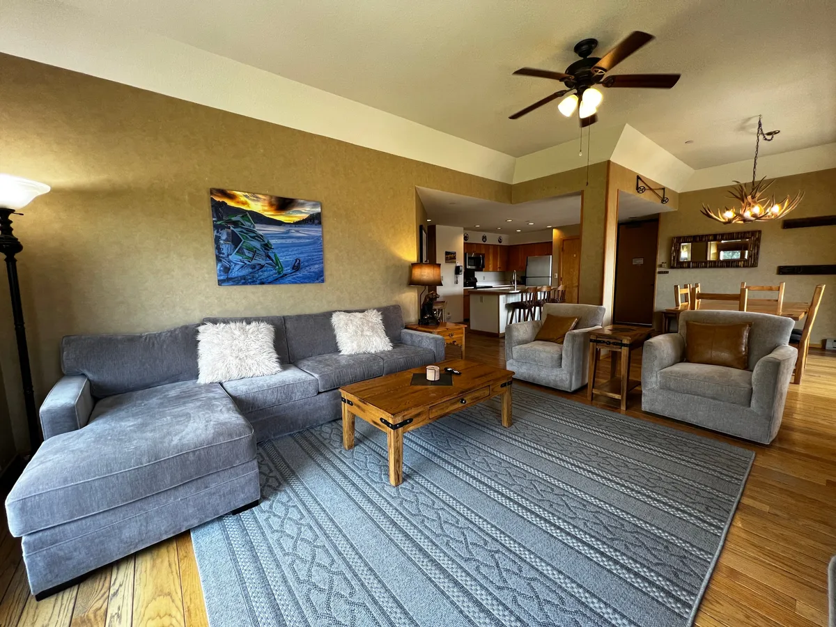 The image showcases a warm, inviting open-concept living space featuring a plush gray sectional sofa, a stylish wooden coffee table, tasteful wall art, ambient lighting from a modern lamp, and a cozy dining area in the background. The decor and layout create a space ideal for gathering, relaxing, and enjoying mountain views after a day on the slopes.
