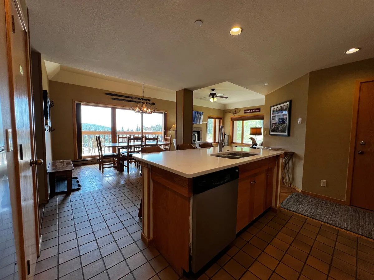 An open-plan area combining a kitchen island, a dining table, and a living space with a fireplace, all illuminated by large windows that reveal sweeping mountain vistas. The scene suggests gatherings, game nights, and a seamless flow between relaxation and social activities—all set against a scenic backdrop.