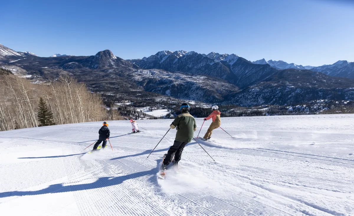 Skiers glide down snow-blanketed slopes with majestic mountains rising behind them, perfectly encapsulating the thrill of winter sports and the natural alpine setting. The scene is dynamic and family-friendly, illustrating active enjoyment of the outdoors.