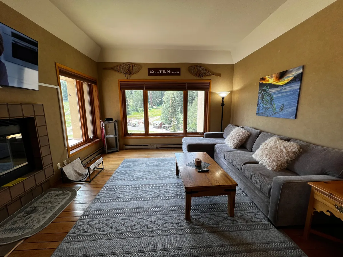 A welcoming living room featuring a comfortable sofa with plush cushions, a wooden coffee table, and expansive floor-to-ceiling windows that frame breathtaking views of the snow-capped mountains. The setting is bathed in warm, natural light, inviting guests to relax, read, or gather with family and friends while taking in the scenery.